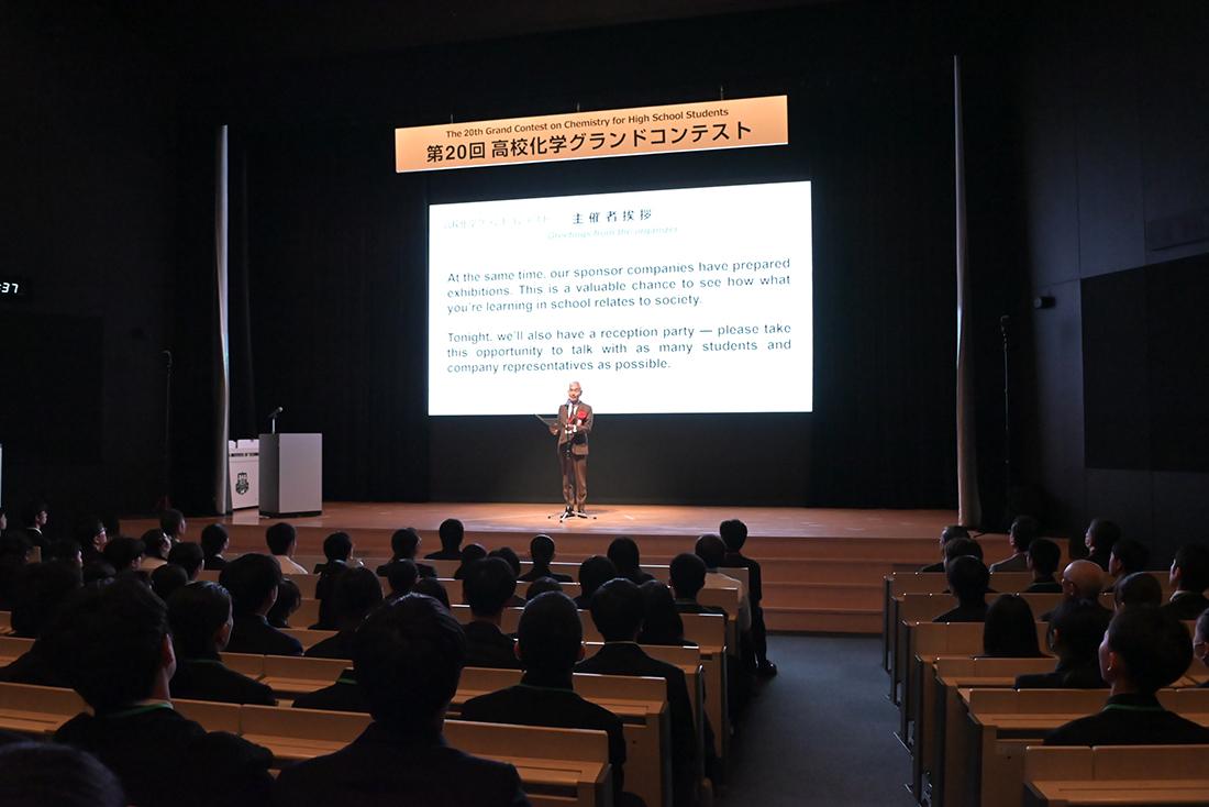 客席が埋まった会場で、壇上の挨拶とスライド投影が行われている 