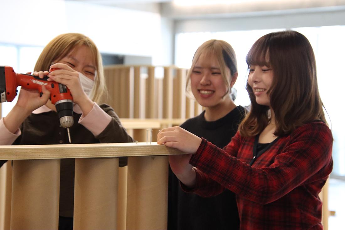 人物が電動ドリルで木製の天板を留め、周囲の2人が支えている