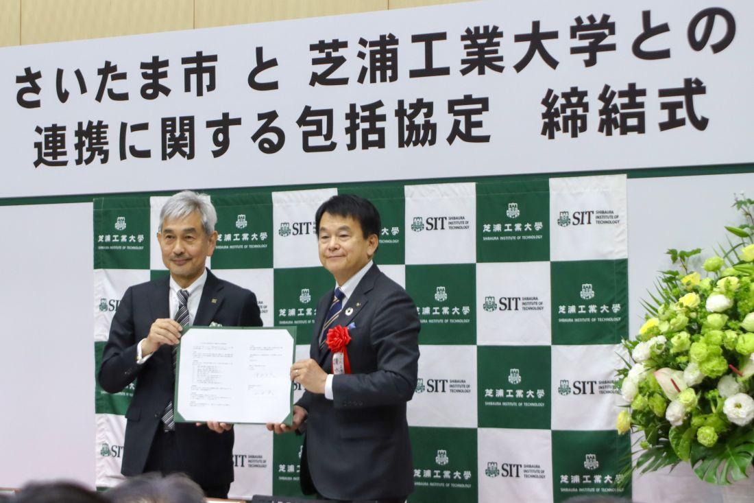 協定締結式の看板前で、2人が署名済みの書類を持って記念撮影している  