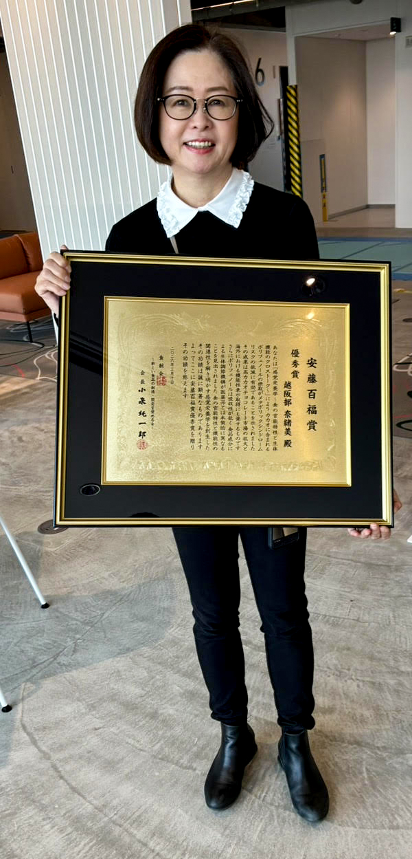 室内で、人物が額装された表彰状（賞状）を胸の前で持って立っている  